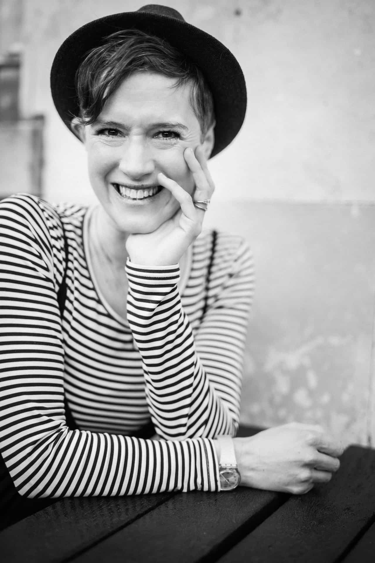 Black and white image of Becca Benning, a white woman with short dark hair, smiling to camera wearing a black and white stripy top and a grey hat - chin resting in her hand.