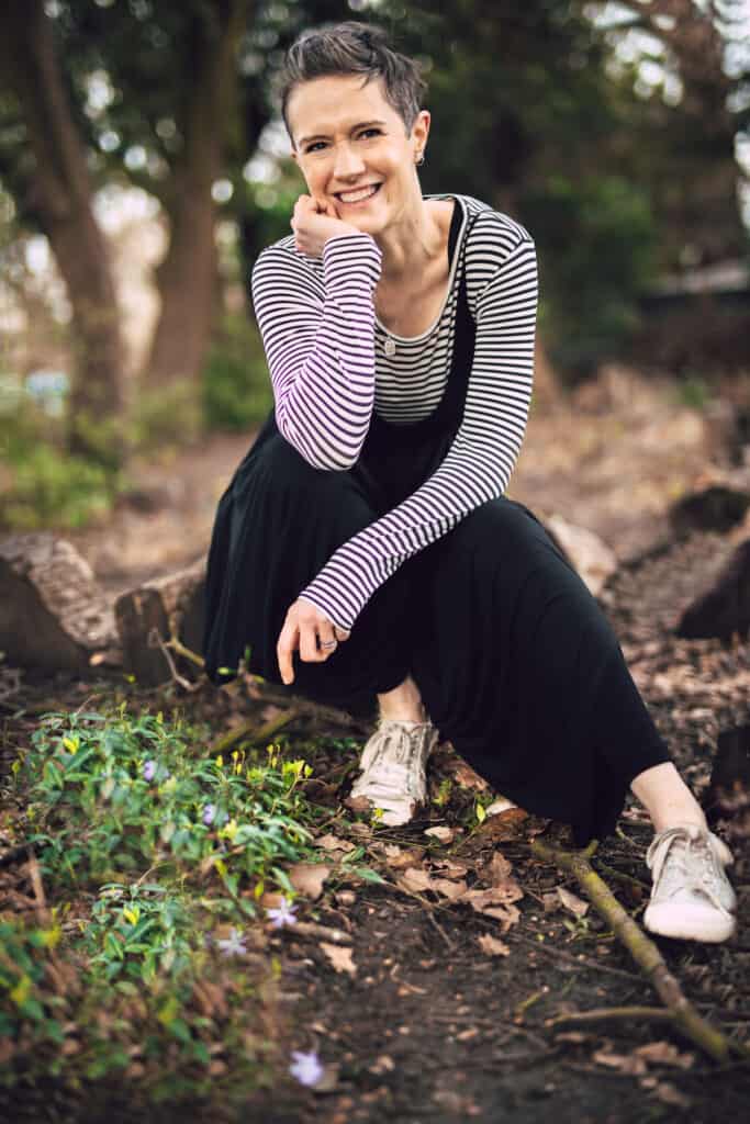 Becca Benning sitting in the woods, smiling. Wearing a loose black jumpsuit with a black and white striped top underneath.