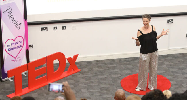 Becca stands on a red dot to the right of the image, with the TEDx sign on the left. Wearing sliver sequinned trousers and a black top, smiling. Some tops of heads in the front of the crowd are visible. Banner to the left shows TEDx Lewisham presents: The Power of Kindness. Talk is called Everyday Ways to Love a Struggling Body.